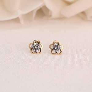 May include: A pair of gold-coloured flower-shaped stud earrings. Each earring has a clear, sparkling centre stone surrounded by five petals. The earrings are set against a soft, off-white background.