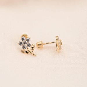 以下が含まれることがあります： 金色の花のスタッドピアス。各イヤリングは、中央に小さなクリアストーンと繊細な葉と茎のディテールを備えた花のデザインが特徴です。イヤリングは、柔らかいオフホワイトの背景に配置されています。