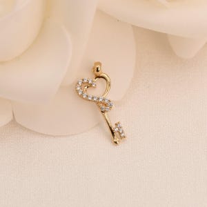 May include: A gold-coloured key pendant with a heart-shaped top, embellished with small, clear gemstones. The key's handle also features a small, gemstone-studded design. The pendant is set against a soft, off-white background.