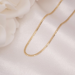May include: A delicate gold chain necklace with a flat curb link design. The necklace is displayed on a white satin surface, with a white flower in the background. The chain is approximately 40 cm long.