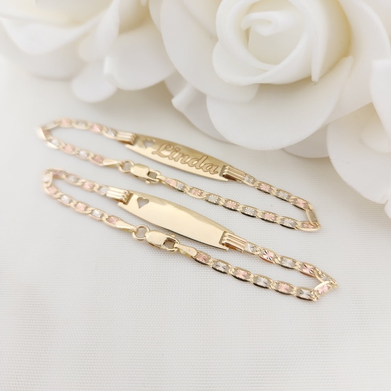 K&ouml;nnte beinhalten: Zwei goldene Armb&auml;nder mit einem Namensschild-Anh&auml;nger. Das obere Armband hat den Namen "Linda" auf dem Anh&auml;nger graviert. Das untere Armband hat einen herzf&ouml;rmigen Ausschnitt auf dem Anh&auml;nger.