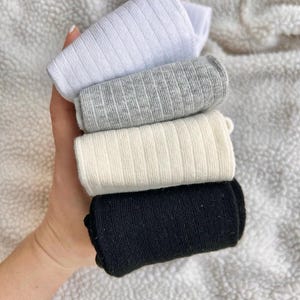 Pode incluir: Uma pilha de quatro meias caneladas dobradas em cores sólidas, incluindo branco, cinza mesclado, creme e preto. Cada meia apresenta uma textura canelada vertical consistente em todo o tecido, disposta em camadas organizadas.