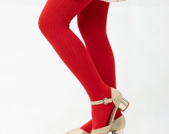 Puede incluir: Medias rojas brillantes con costuras acanaladas combinadas con tacones Mary Jane dorados con purpurina. Los tacones tienen un tacón de bloque pequeño y un cierre de hebilla. El conjunto se completa con una falda con estampado floral.