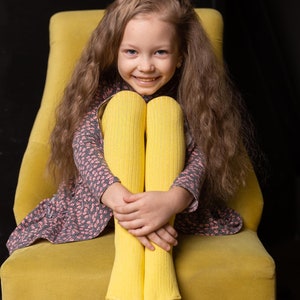 May include: A young girl with long brown hair sits in a yellow armchair. She is wearing a pink and grey dress and yellow tights. She is smiling at the camera.