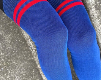 May include: A pair of over-the-knee socks in a vibrant royal blue color. The socks feature a ribbed cuff at the top with two horizontal red stripes. The fabric appears to be a knit material, extending from the upper thigh down to the lower leg area.
