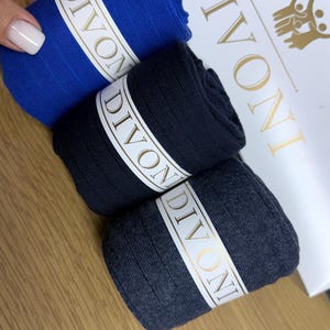 May include: Three pairs of ribbed socks in blue, black, and dark grey, each featuring a white band with the text DIVONI printed in gold. The socks are positioned in front of a white box with the brand name DIVONI and a gold logo printed on it.