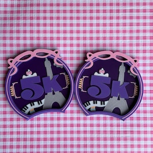 May include: Two purple and pink 5K race medallions with a Parisian theme. Each medallion features the text "5K", an Eiffel Tower silhouette, and a piano keyboard design. The background is a pink and white checkered pattern.