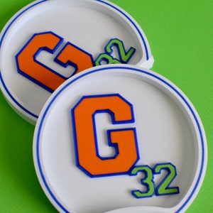 May include: Two white coasters with blue trim. The coasters feature a large orange letter "G" with a blue outline. The number "32" is also on the coasters in green with a white outline.