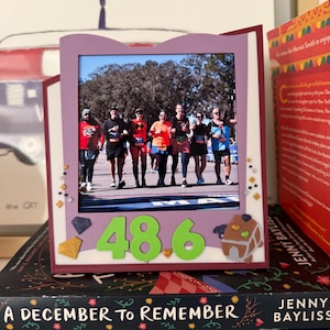 May include: A framed photo featuring a group of people running a race. The frame is purple with decorative embellishments. The number "48.6" is displayed in green, along with a gingerbread house and a diamond. The frame rests on a stack of books.