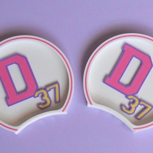 May include: A pair of white mouse ears with a pink and purple "D" and the number "37" in yellow on each ear.