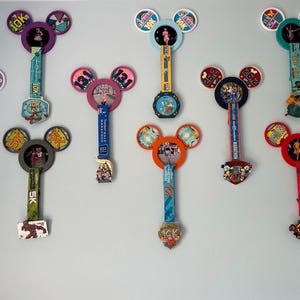 May include: A collection of nine colourful Disney race medals, each featuring a Mickey Mouse ear design with a different race distance and theme. The medals are displayed on a wall, with the ribbons hanging down.