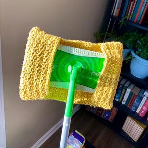May include: A yellow crocheted cover for a Swiffer Sweeper mop head. The cover is crocheted in a simple stitch and has a slightly textured look. The mop head is green and has the word "Swiffer" printed on it.
