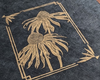 May include: A black slate coaster with a gold foil design of two stylized flowers in a frame. The flowers are in a vintage art deco style.
