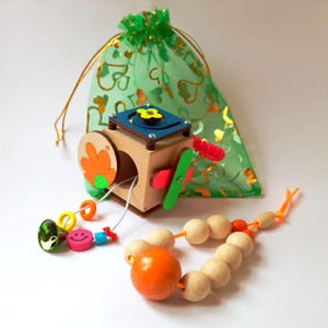 May include: A colorful wooden toy with a variety of textures and activities for babies and toddlers. The toy includes a cube with moving parts, a bead necklace, and a small bell. The toy is packaged in a green organza bag.