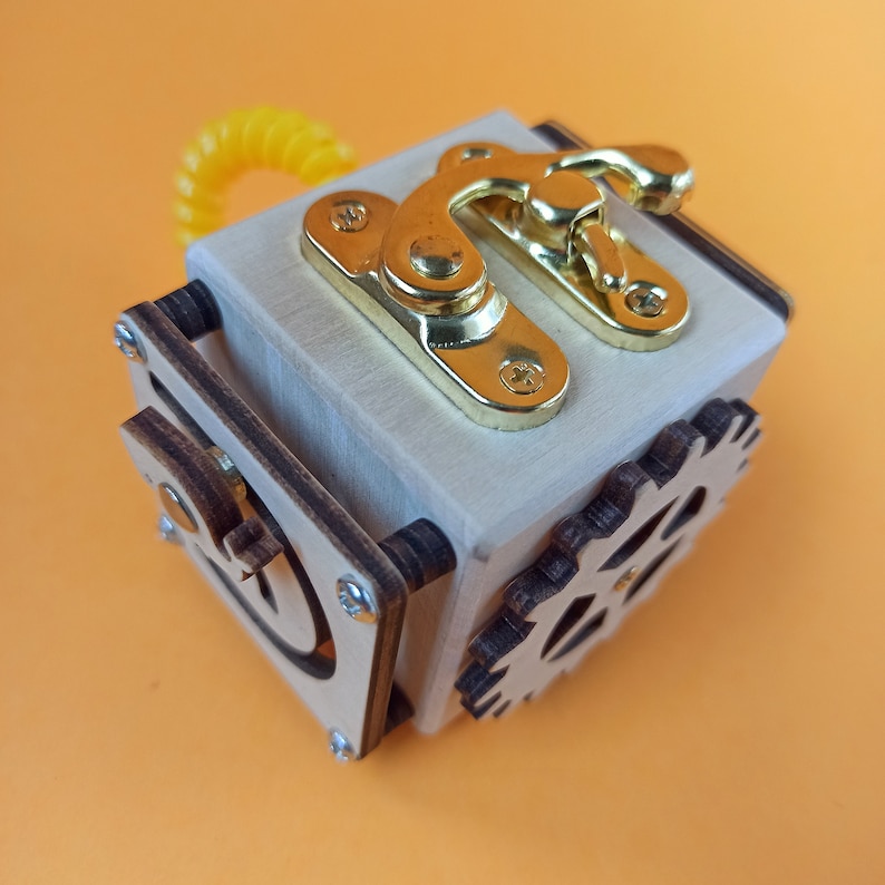 May include: A wooden box with a gold latch and a gear on the side. The box is made of light wood and has a dark wood trim.
