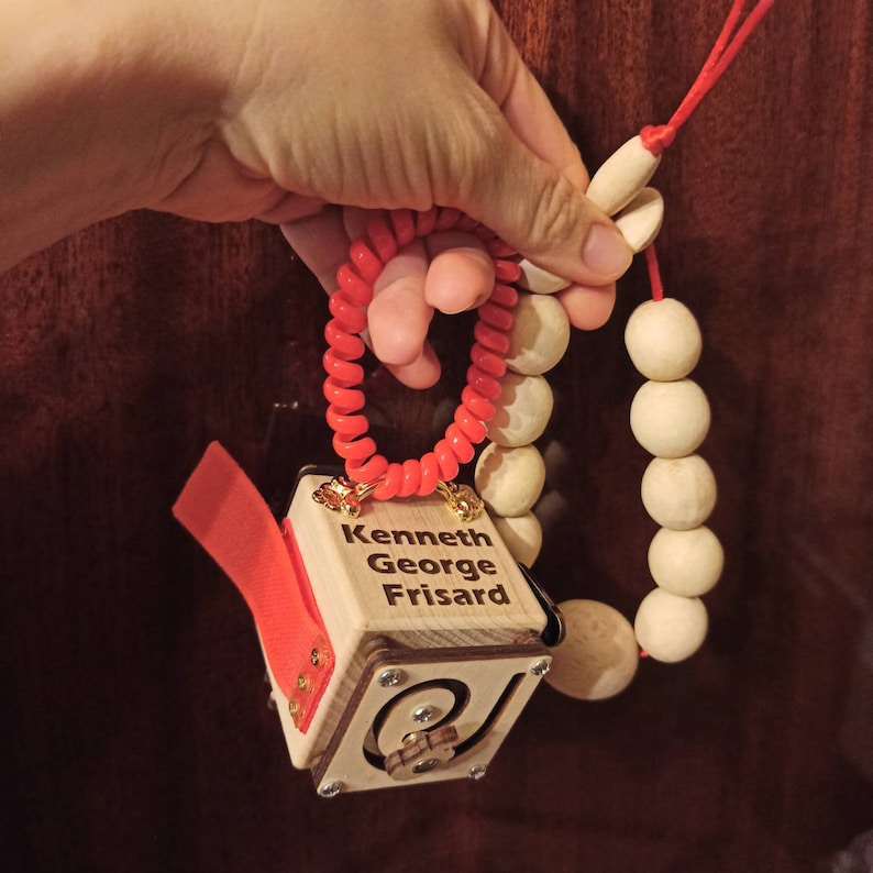 May include: A wooden cube with the name "Kenneth George Frisard" engraved on it. The cube is attached to a wooden bead necklace with a red spiral hair tie.