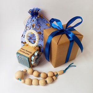 Puede incluir: Una colección de regalos, que incluye un bloque de madera con el texto "Your family loves you", una caja envuelta con una cinta azul, una bolsa de organza azul y un collar de cuentas de madera. El bloque tiene un asa blanca y detalles azules.