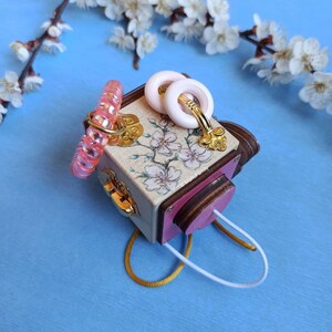 May include: A small wooden box with a pink and white floral design. The box has a gold latch and is decorated with pink and white hair ties and a pair of pink and white earrings.