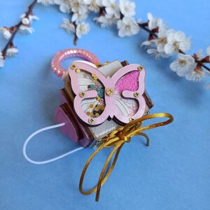 May include: A pink wooden box with a butterfly design. The butterfly has pink and white wings with glitter and beads. The box has a gold ribbon and a white cord.