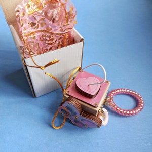 May include: A wooden box with a pink heart cutout and a butterfly design. The box is attached to a pink beaded bracelet and a white string. The box is sitting on a blue surface.