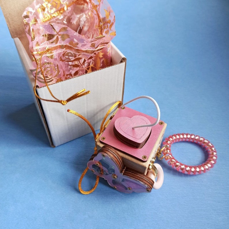May include: A wooden box with a pink heart cutout and a butterfly design. The box is attached to a pink beaded bracelet and a white string. The box is sitting on a blue surface.