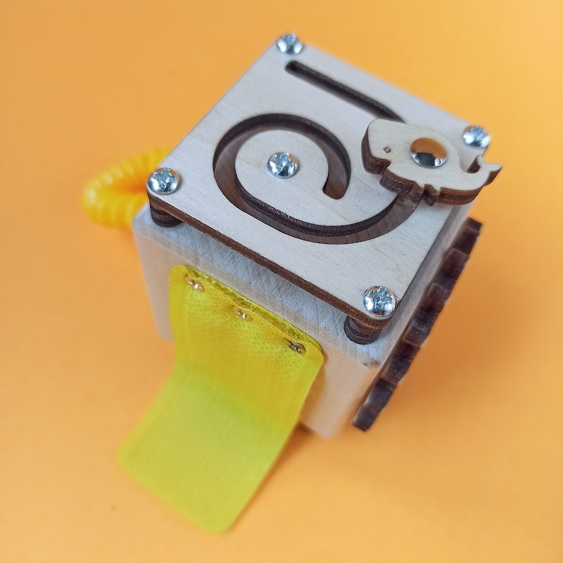 May include: Wooden toy cube with a yellow fabric flap and a maze on the top. The maze has a small wooden dinosaur figure that can be moved around the maze.