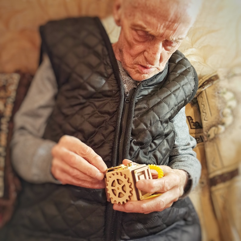 May include: A person is holding a wooden puzzle box with gears and a yellow rubber band. The box is made of light brown wood and has a intricate design.