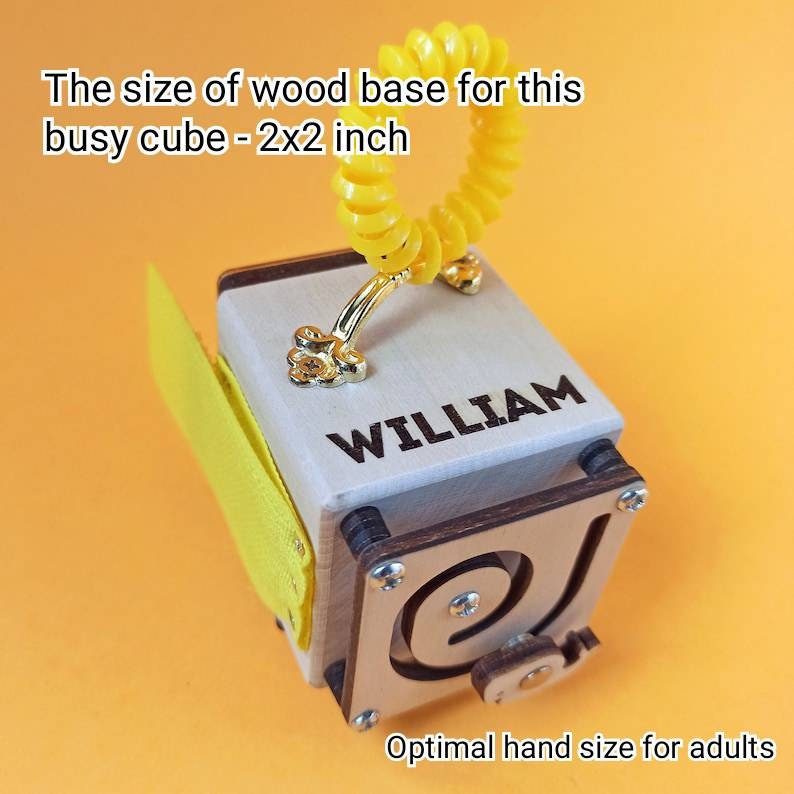 May include: A wooden busy cube with a yellow fabric flap, a gold handle, and a spiral toy. The cube has a wooden base that measures 2 inches by 2 inches. The cube is engraved with the name "WILLIAM".