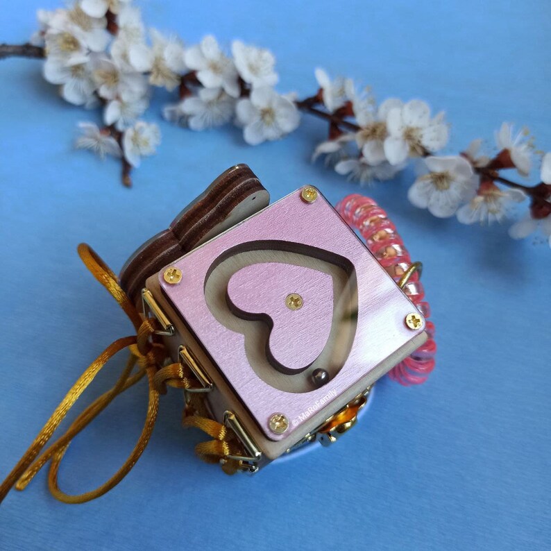 May include: A pink wooden box with a heart-shaped maze and a small metal ball inside. The box is decorated with gold trim and a gold ribbon. The box is sitting on a blue surface.