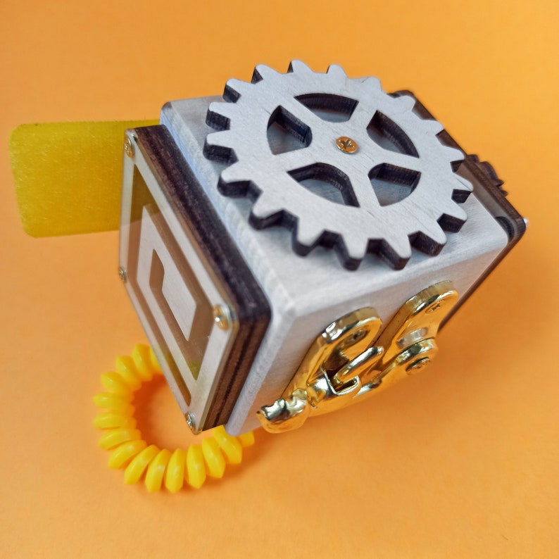 May include: A wooden toy with a white gear, gold metal accents, and a yellow rubber band. The toy has a square shape with a clear plastic window showing a geometric design.