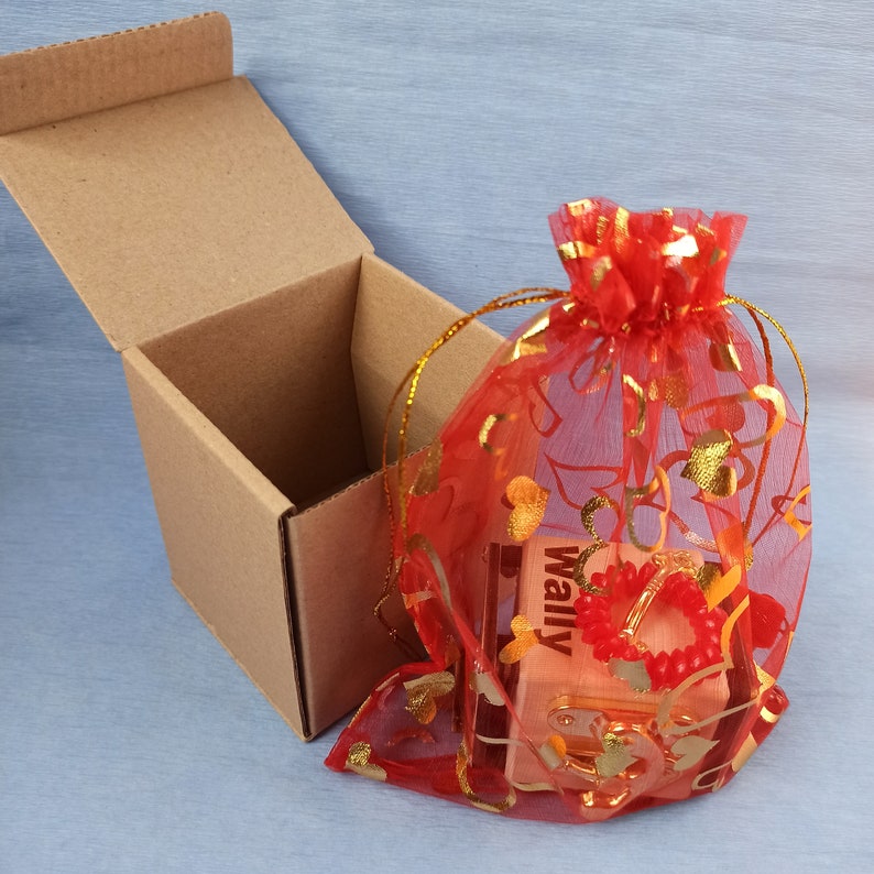 May include: A small wooden box with the word "Wally" printed on it, wrapped in a red sheer bag with gold hearts and floral designs. The box is sitting inside a brown cardboard box.