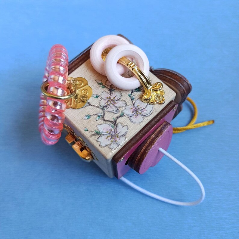 May include: A small wooden box with a pink and white floral design. The box has a gold handle and a pink and white ribbon. The box is decorated with a pink and white floral design. The box is decorated with a pink and white floral design.