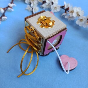 May include: A wooden cube toy with a pink heart-shaped panel, a gold latch, and a yellow cord. The cord is threaded through the cube and tied in a bow. The cube is sitting on a blue surface.