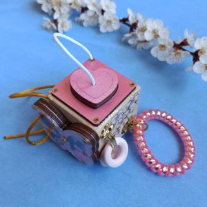 May include: A wooden sensory cube with a pink heart, a butterfly, a flower design, and a pink spiral. The cube has a white string loop and a pink ring.