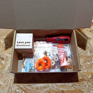 Könnte beinhalten: Ein offener Pappkarton gefüllt mit verschiedenen Gegenständen. Ein Holzblock mit der Gravur "Love you Grandma" befindet sich neben einem roten Schraubendreher. Weitere Artikel sind orangefarbene Accessoires, kleine Zahnräder und verpackte Hardware.