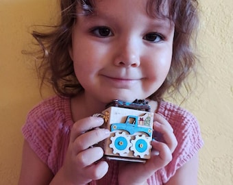 May include: A small wooden cube toy featuring a light blue truck graphic with a Christmas tree in the bed, two white gear-shaped wheels attached with gold screws, and a natural wood finish on the sides, designed for tactile sensory play.