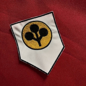 May include: A white patch with a black border and a gold circle containing a black floral design. The patch is set against a red background.