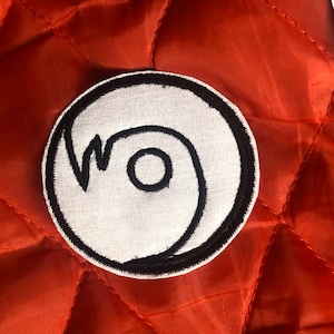 Op de afbeelding: Witte stoffen patch met een zwarte omtrek van een gestileerd swirl-ontwerp. De patch is rond en heeft een zwarte rand.