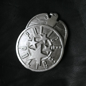 May include: A silver-colored police badge with the word "POLICE" encircling a star and three smaller stars. The badge is set against a black leather-like background.