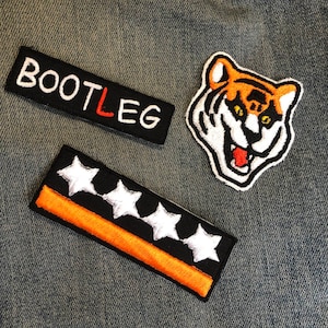 Puede incluir: Tres parches bordados sobre un fondo de mezclilla. Los parches son negros con costuras blancas y presentan las palabras "BOOTLEG", cuatro estrellas blancas y una cabeza de tigre de dibujos animados con rayas naranjas y negras.