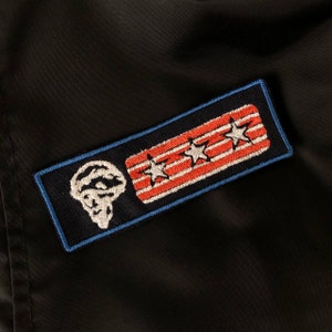 Puede incluir: Un parche bordado en negro y azul con una calavera blanca y tres estrellas blancas sobre un fondo a rayas rojo y blanco.
