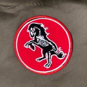 Pode incluir: Um remendo circular vermelho e branco com uma silhueta preta de um esqueleto de cavalo empinado. O cavalo está voltado para o lado esquerdo da imagem.