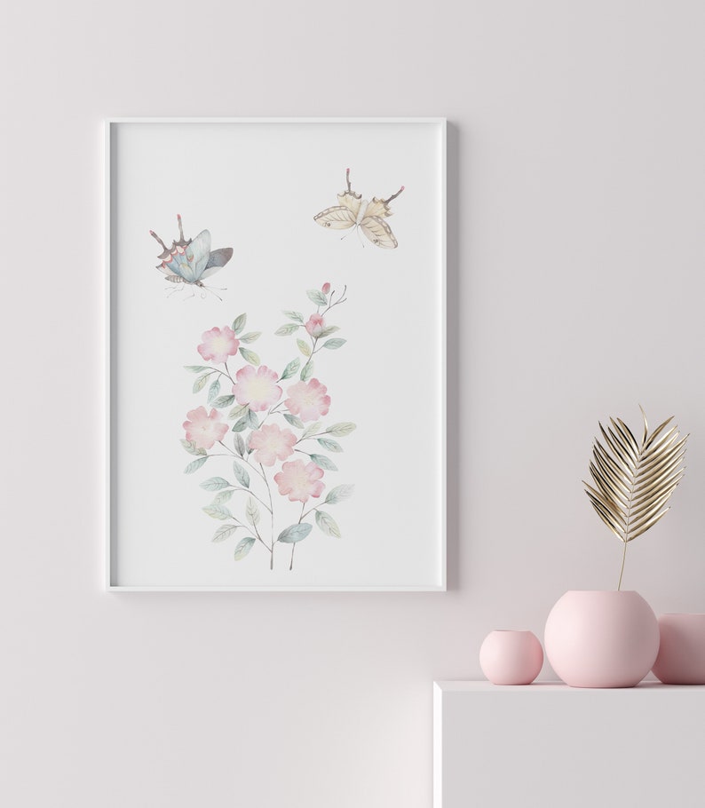 Op de afbeelding: Een ingelijste aquarelprint met roze bloemen en twee vlinders. De vlinders hebben blauwe en bruine vleugels. De bloemen hebben groene bladeren en stelen. De print zit in een witte lijst.