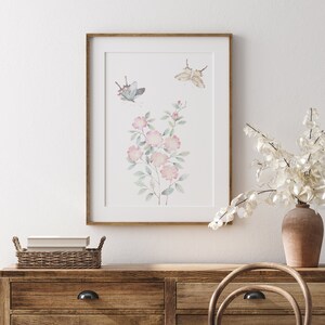 Op de afbeelding: Een ingelijste aquarelprint met een bloemstuk met roze bloesems en groene bladeren, samen met twee vlinders. Het kunstwerk is geplaatst tegen een witte achtergrond en ingelijst in een lichtbruine houten lijst. De print wordt getoond op een houten dressoir.