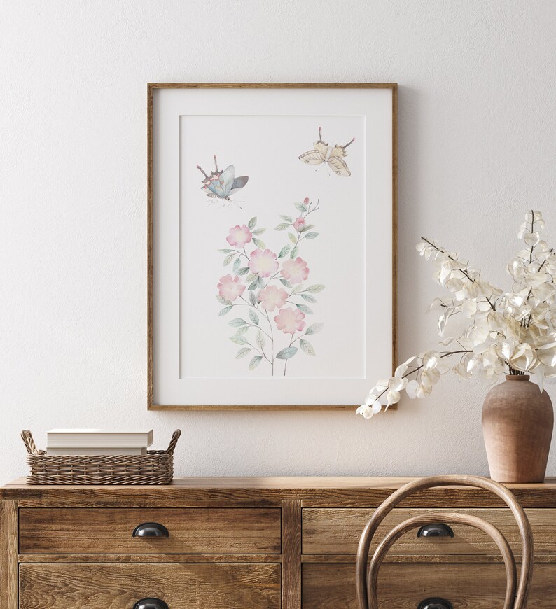 Op de afbeelding: Een ingelijste aquarelprint met een bloemstuk met roze bloesems en groene bladeren, samen met twee vlinders. Het kunstwerk is geplaatst tegen een witte achtergrond en ingelijst in een lichtbruine houten lijst. De print wordt getoond op een houten dressoir.