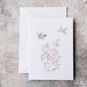 Op de afbeelding: Een witte wenskaart met een aquarel illustratie van roze bloemen en twee vlinders. Een bijpassende witte envelop bevindt zich achter de kaart. Het ontwerp van de kaart heeft een bloemen- en vlinderthema.
