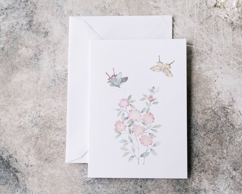 Op de afbeelding: Een witte wenskaart met een aquarel illustratie van roze bloemen en twee vlinders. Een bijpassende witte envelop bevindt zich achter de kaart. Het ontwerp van de kaart heeft een bloemen- en vlinderthema.