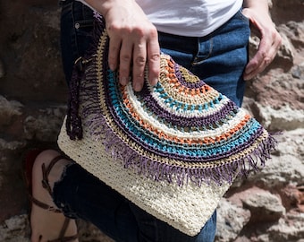 May include: A crocheted clutch bag with a fringe detail. The bag is made with a variety of colors, including white, brown, orange, blue, and purple. The bag is held by a person wearing blue jeans.