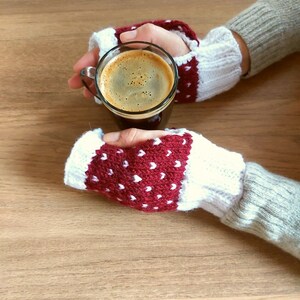 May include: A pair of red and white fingerless gloves with white polka dots, held by hands holding a glass mug of coffee.