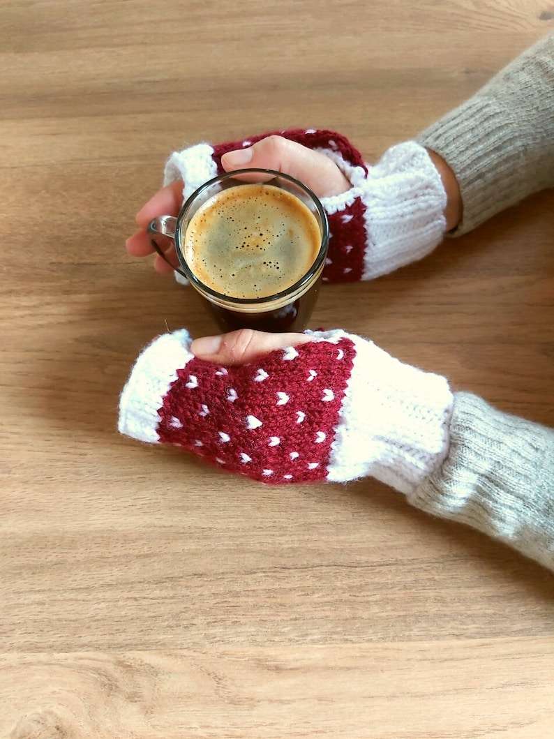 May include: A pair of red and white fingerless gloves with white polka dots, held by hands holding a glass mug of coffee.